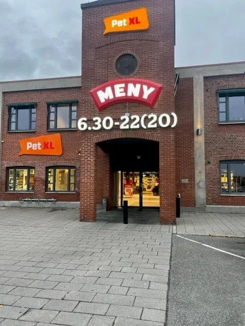Inngangsparti til MENY og Pet XL i mursteinsbygg, med opplyst skilt og åpningstider over døra.