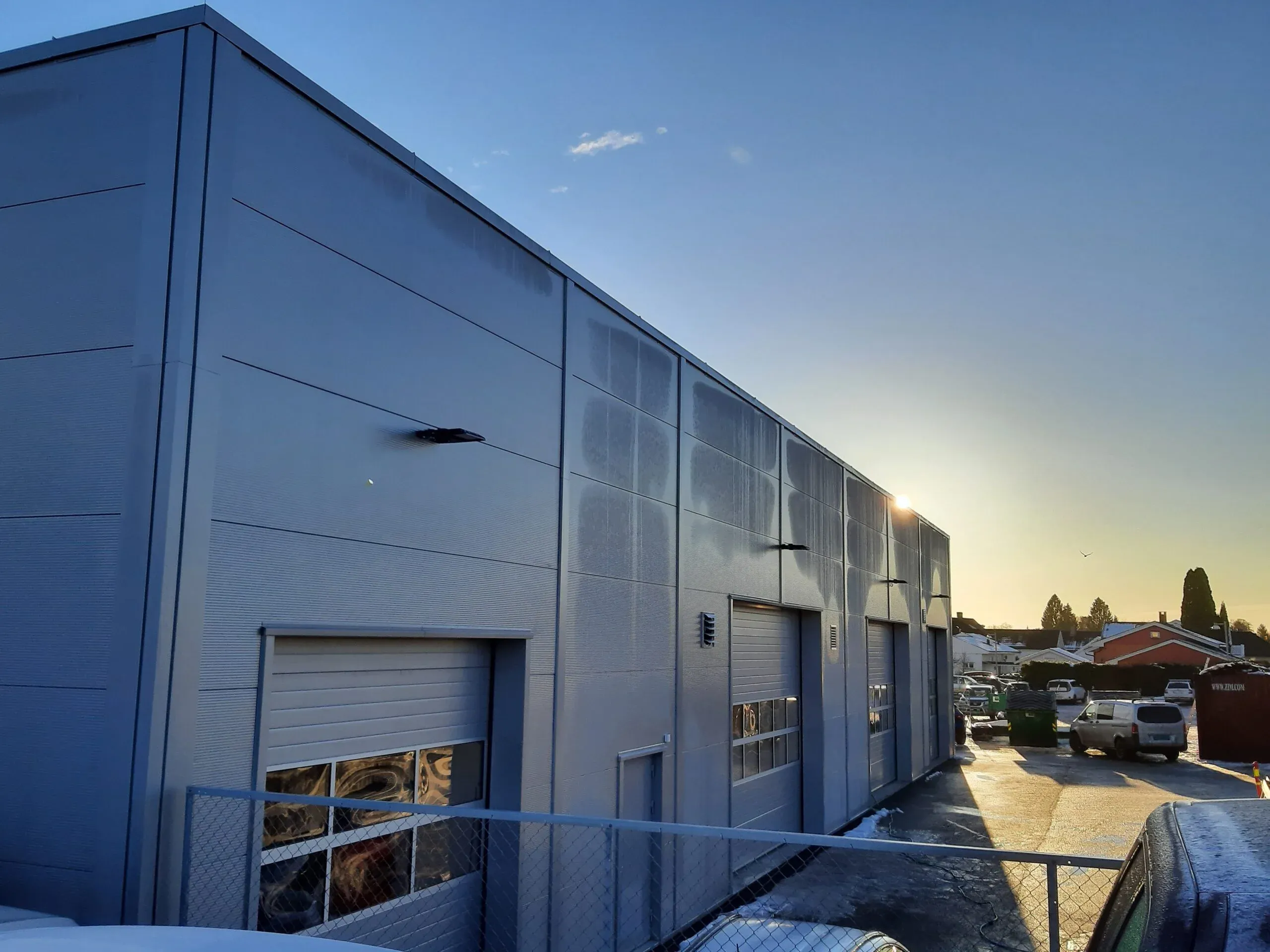 Industribygg med flere garasjeporter og våt asfalt i kveldssol, omgitt av biler og gjerde.