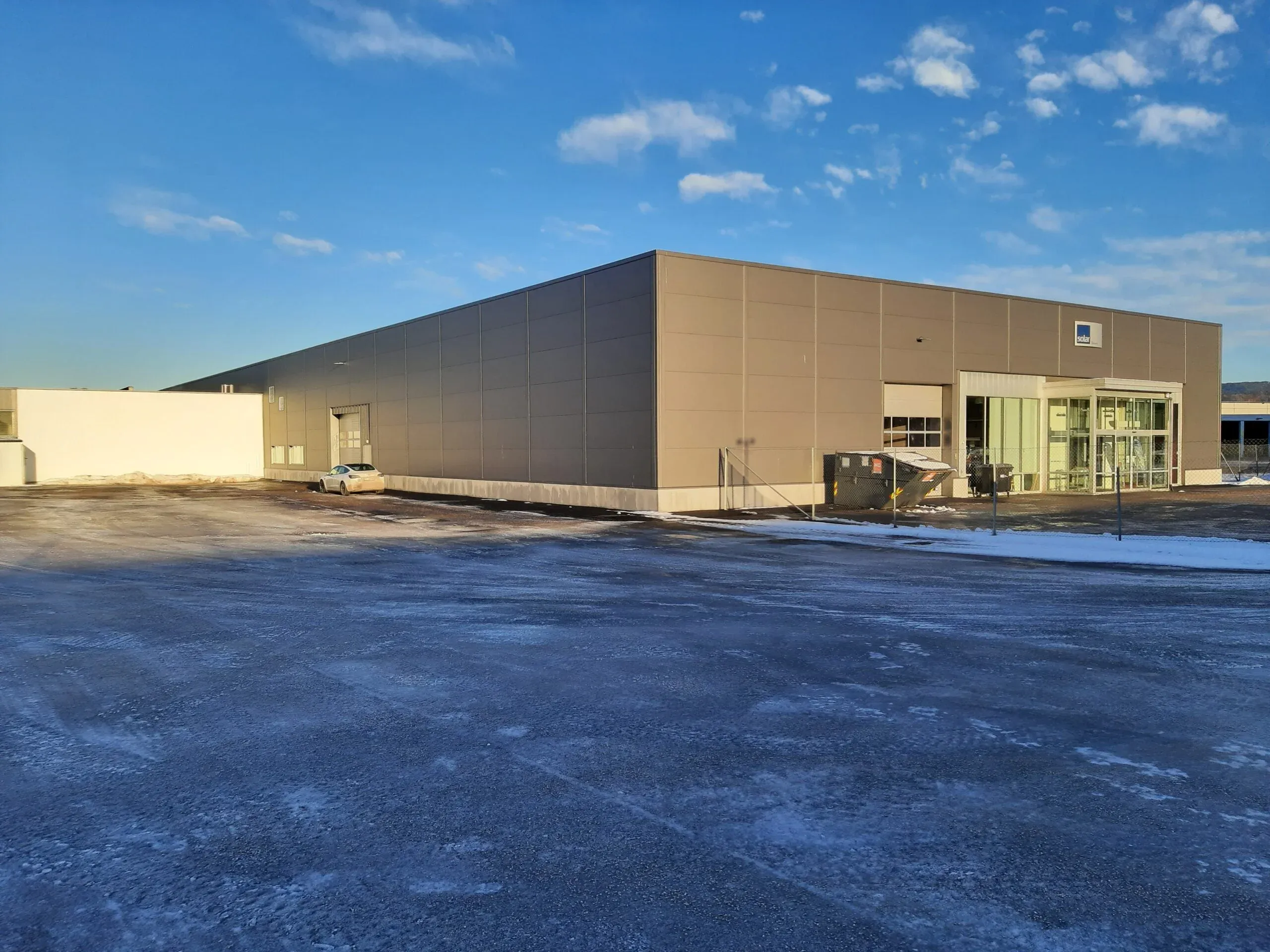 Bredt industribygg med inngangsparti merket "Solar", sett fra siden med stor, snødekket parkeringsplass.