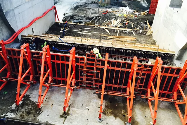 Arbeidere monterer armering og forskaling foran rød støpeform i betongkonstruksjon under bygging.