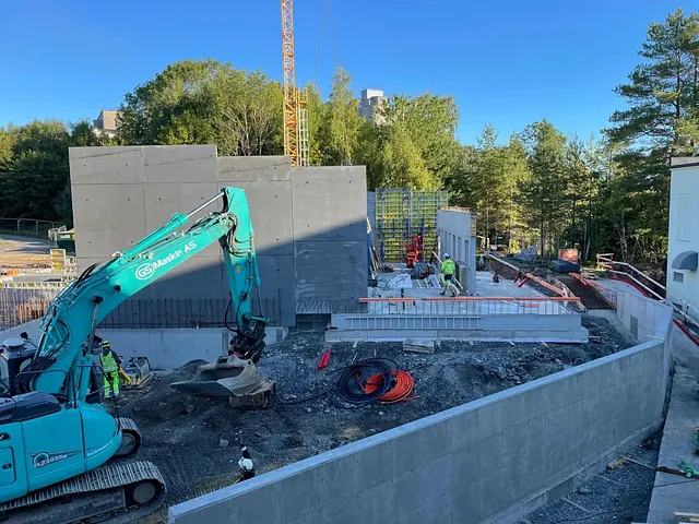 Grønn gravemaskin og arbeidere ved nybygde betongvegger på anleggsområde omgitt av skog.