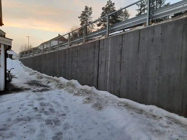 Høy betongmur langs gangsti dekket av snø, med autovern og hus i bakgrunnen under vinterhimmel.