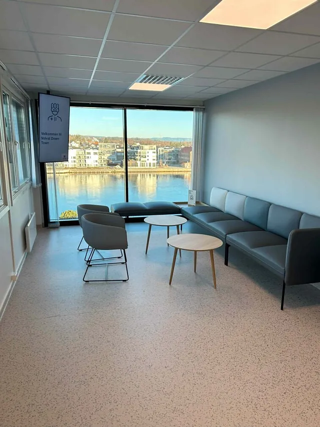 Venterom med store vinduer og utsikt over elven, møblert med grå sofaer, stoler og runde bord.