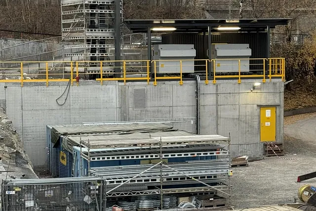 Betongbygg med gult rekkverk og teknisk utstyr på taket, omgitt av stillasdeler og containere på bakken.