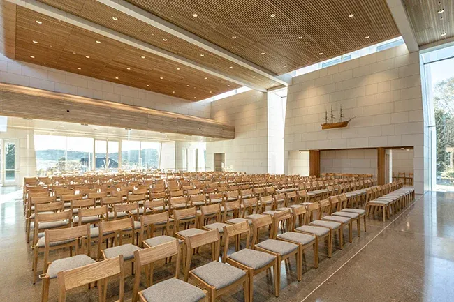 Stort kirkerom med høyt tak, mange lyse trestoler og moderne arkitektur med lysinnslipp.