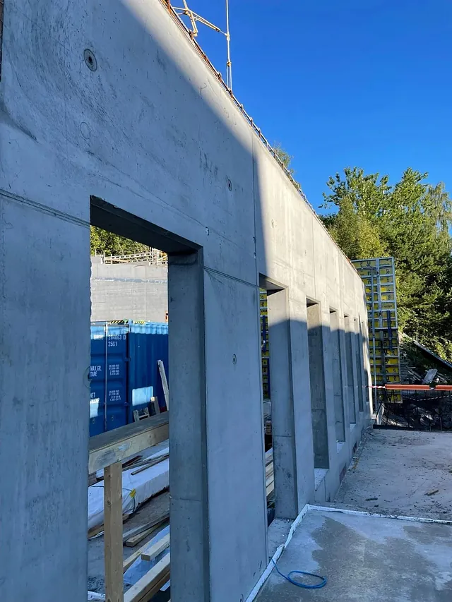 Rett betongvegg med åpninger for dører eller vinduer under bygging, sett fra innsiden av tomta.