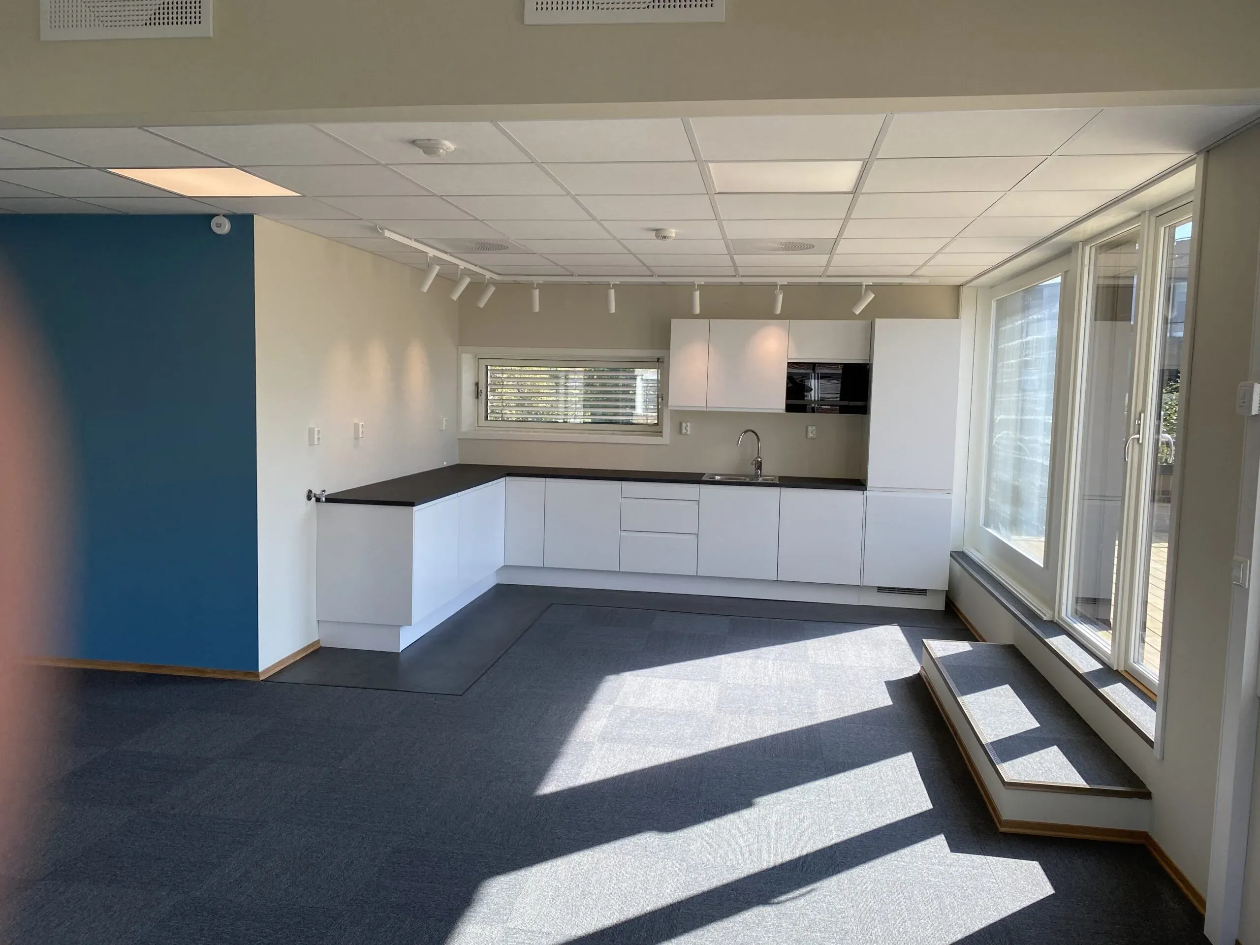 Åpen kjøkkenløsning i kontorlandskap, med moderne hvite skap, grå benkeplate og store vindusflater med dagslys.