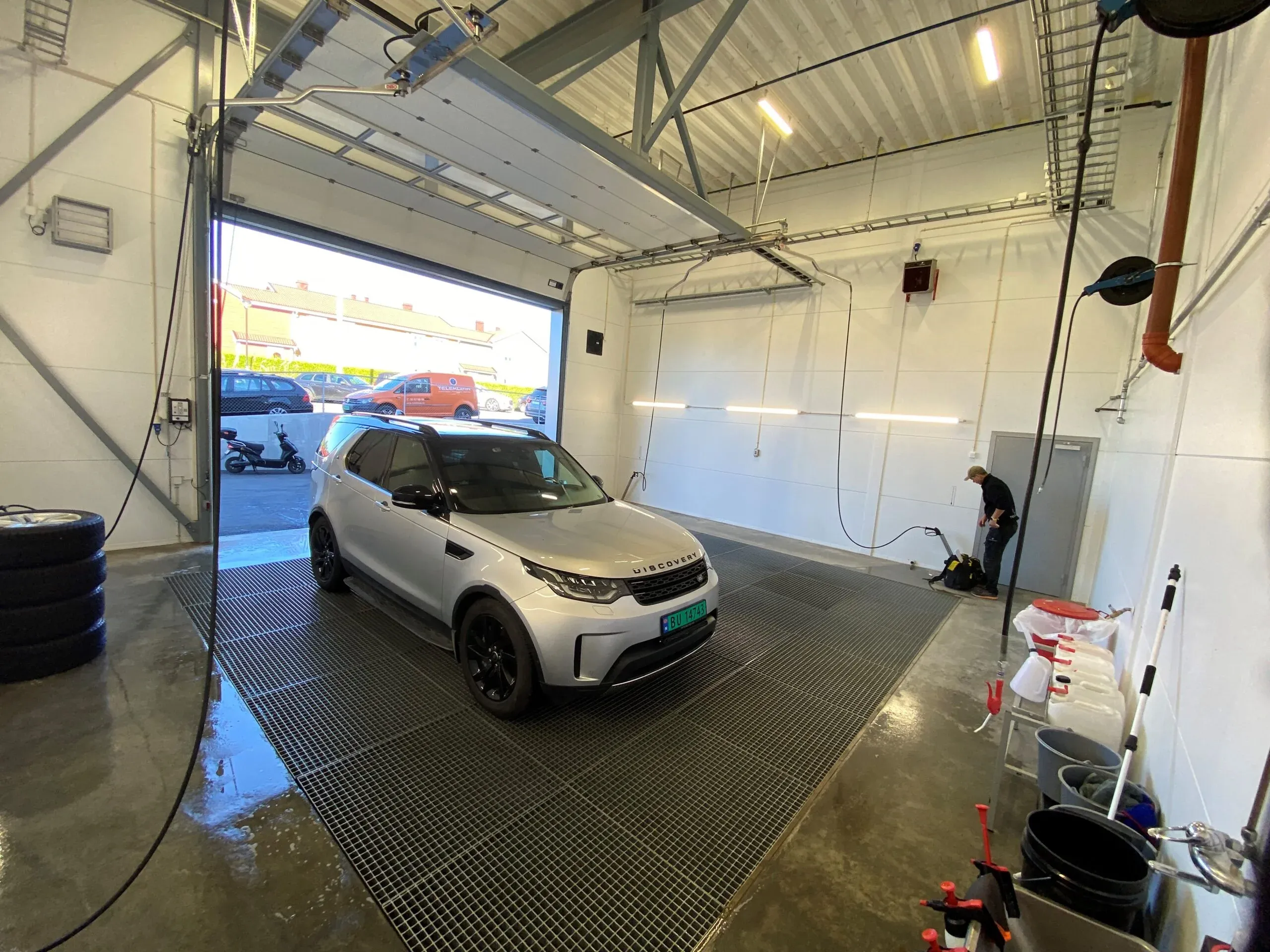 Bilvaskehall med en sølvfarget SUV og utstyr for rengjøring, åpen port ut mot parkering.