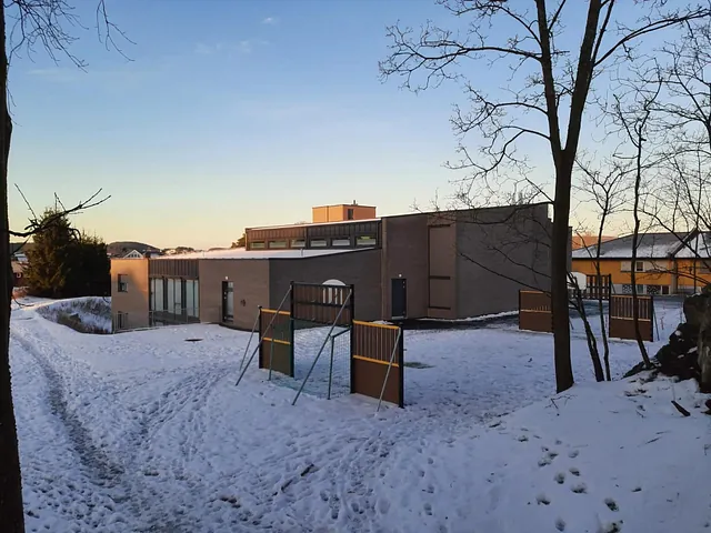 Baksiden av kirkebygget med snødekket lekeområde og trær i forgrunnen.