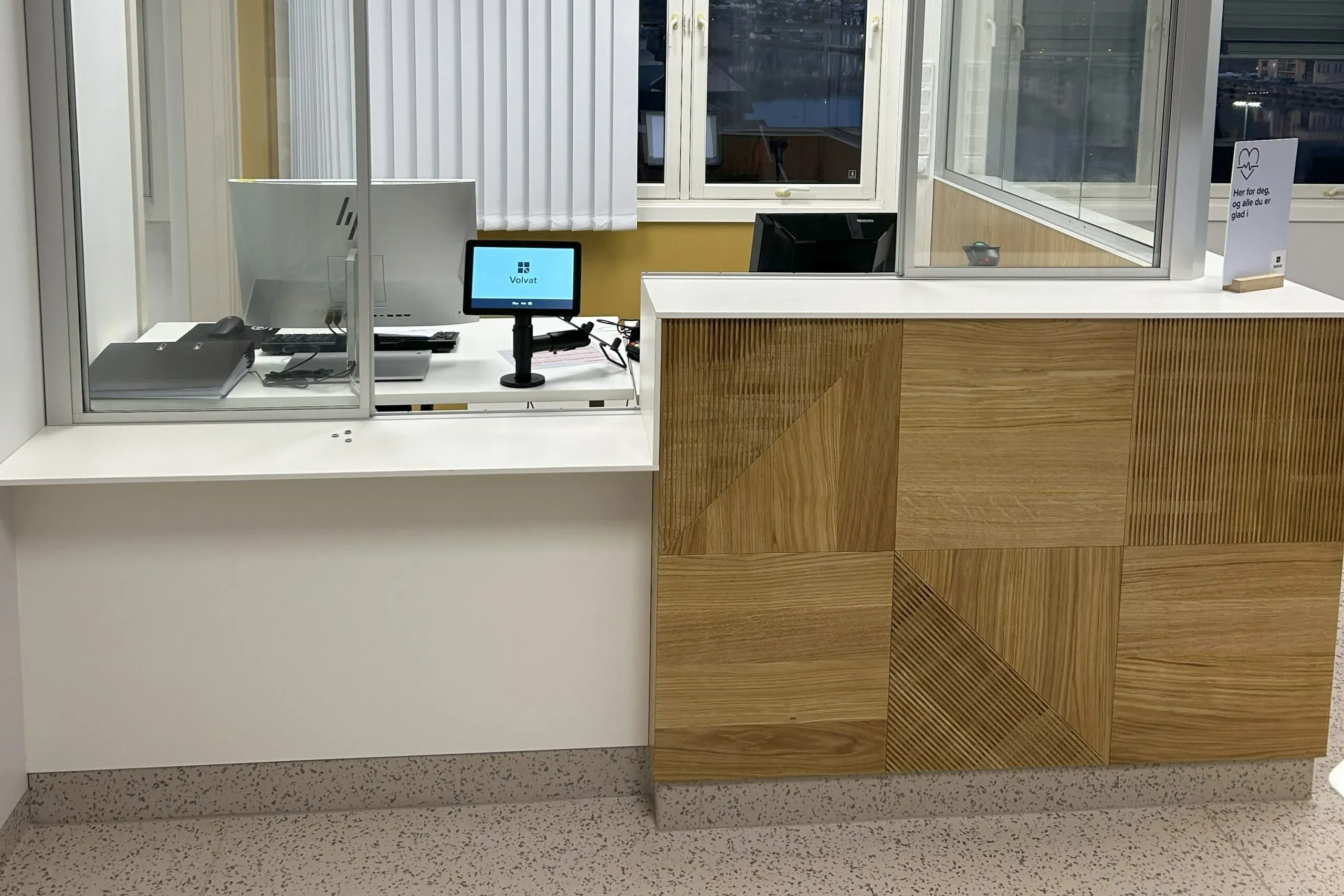 Moderne resepsjon med tredetaljer, skjerm og glassvegg mot kontorområde.