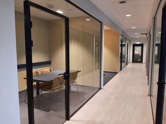Møterom med glassvegg, mørkt bord og gule stoler langs gang i moderne kontorbygg.