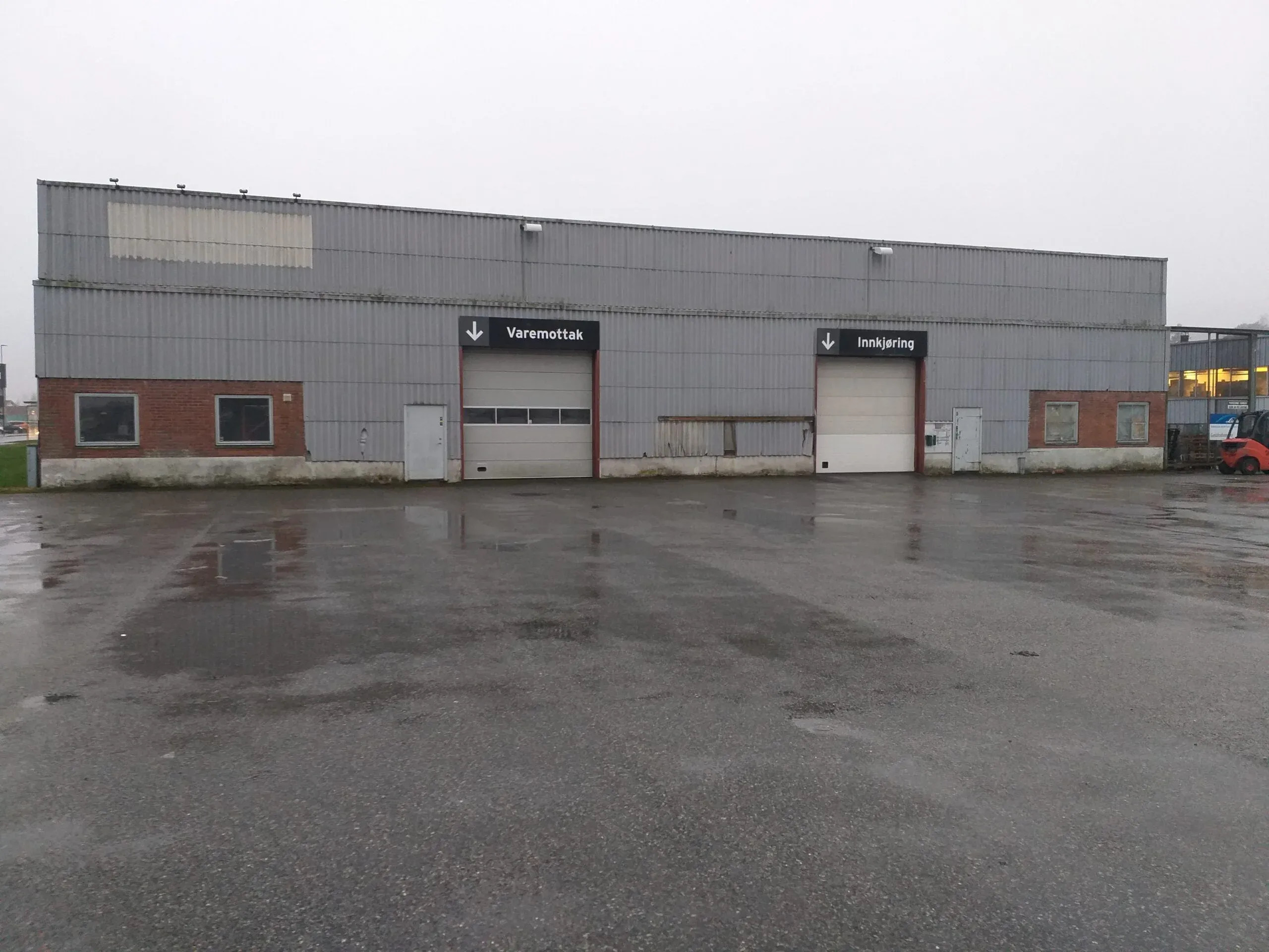 Enkelt industribygg med bølgeblikk og mur, to porter merket "Varemottak" og "Innkjøring" på våt asfalt.
