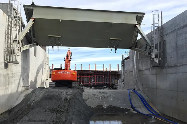 Gravemaskin fra Hitachi i betongkledd kanal, under stor metallport og foran støpearbeid.