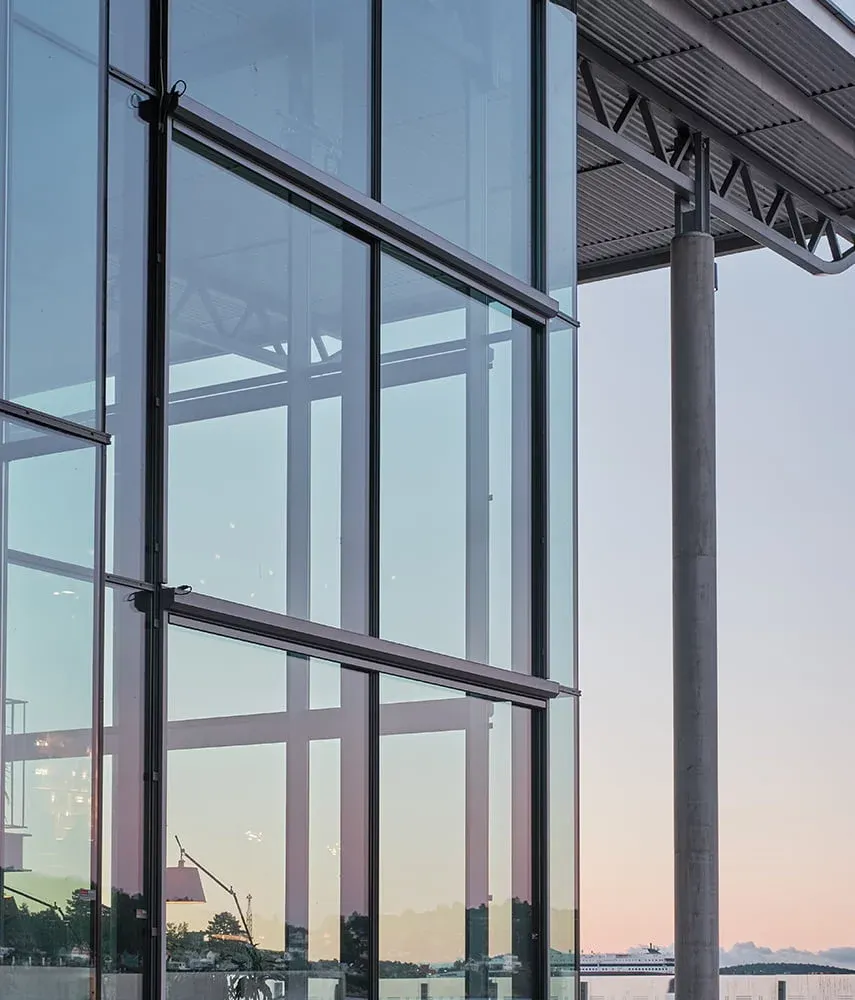 Nærbilde av hjørne med store glassvinduer som reflekterer himmel og landskap i solnedgang.