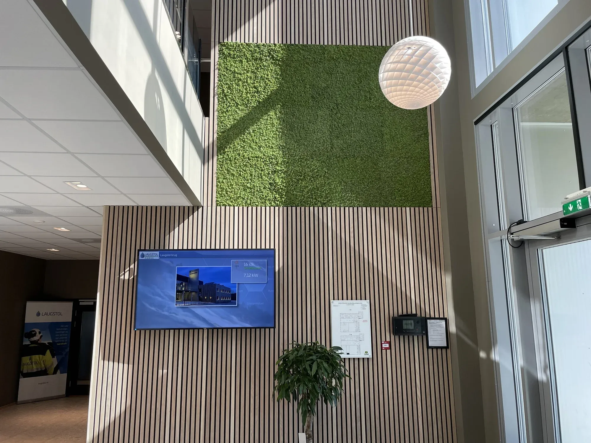 Lyst inngangsparti med grønt mosefelt og informasjonsskjerm montert på vegg med trepanel.