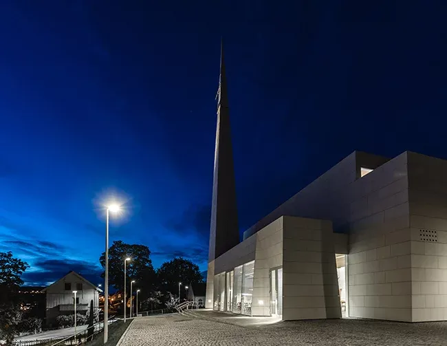 Kirke med moderne hvit fasade og høy spiss tårnspiss, opplyst mot en mørk kveldshimmel.