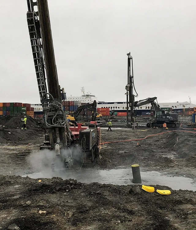 Boreutstyr og gravemaskiner i arbeid på en gjørmete anleggsplass med containere og skip i bakgrunnen.