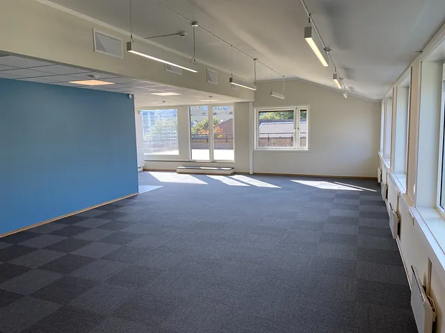 Lyst møterom eller flerbruksrom med blå aksentvegg, grå teppefliser og store vinduer som slipper inn naturlig lys.