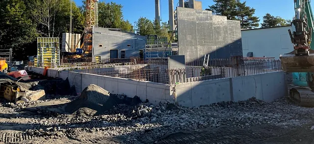 Byggeplass med betongfundamenter og forskaling under montering, omgitt av grus og anleggsmaskiner.