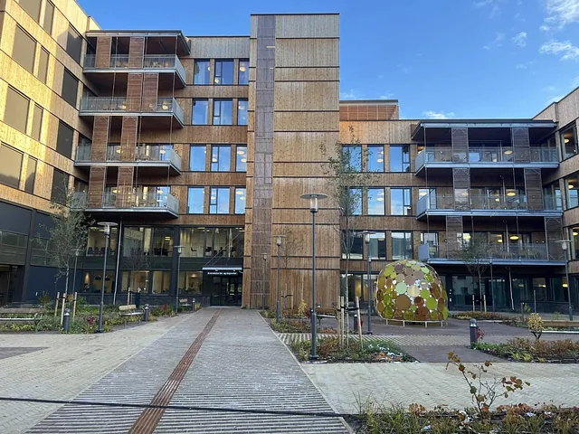 Inngangsparti til nytt bygg med balkonger i tre, omgitt av beplantet gårdsrom og kunstinstallasjon.