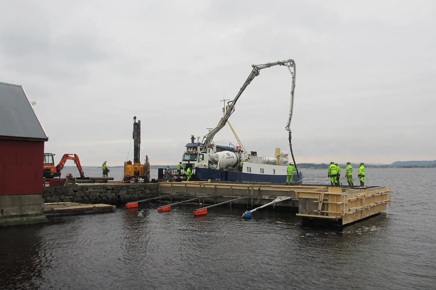 Arbeidere støper kai med flytende forskaling mens betong pumpes fra båt med lang armkran.