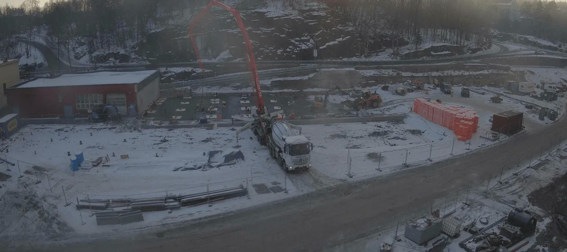Snødekt byggeplass med betongbil, kranarm og anleggsmaskiner i drift, omgitt av materialer og brakker.