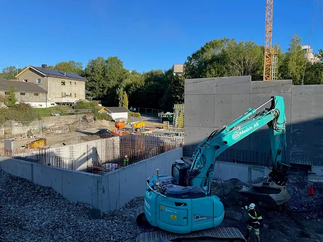 Gravemaskin og arbeidere i sving ved betongkonstruksjon, med bolig og stillas i bakgrunnen.