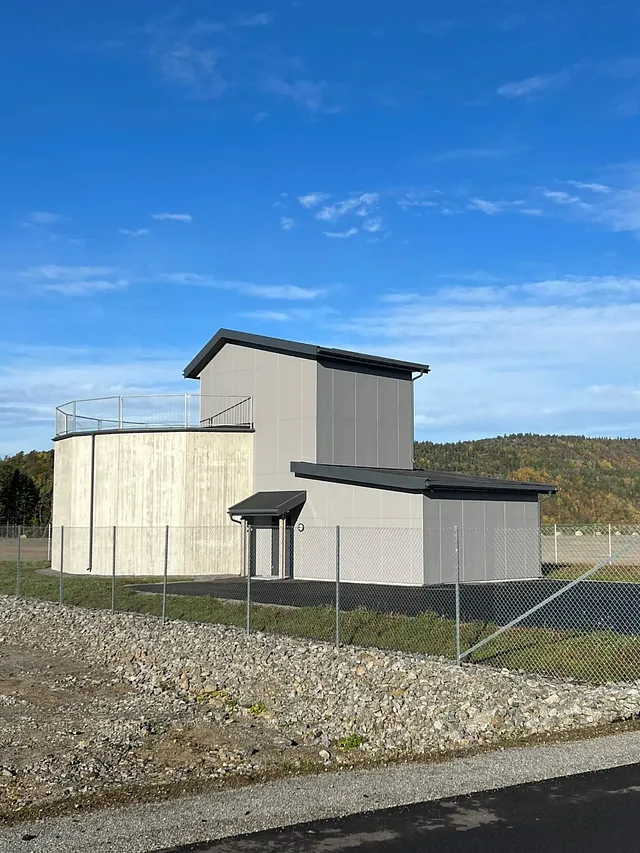 Fasade av teknisk bygg med sylinderformet betongtank, stålgjerde og blå himmel.