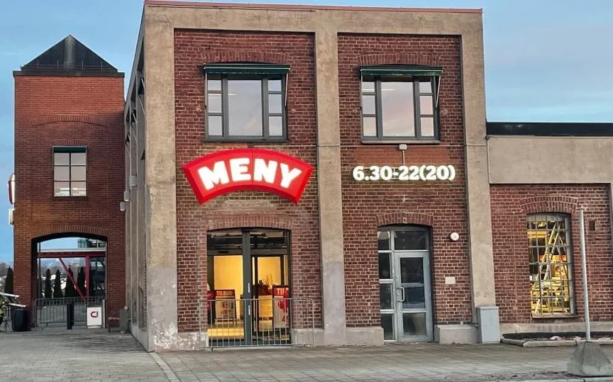 Fasade av MENY-butikk i murbygning med opplyst rødt skilt og åpningstider projisert på veggen.